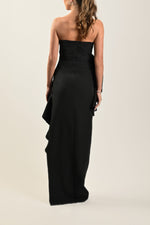 A model wearing a long black dress with a flower detail on the chest and a strapless neckline.